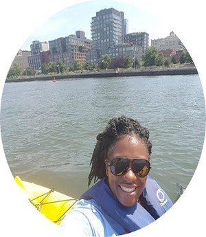 A woman dressed in blue vest and white leeves smiles into camera while a city and river sets in the backdrop 