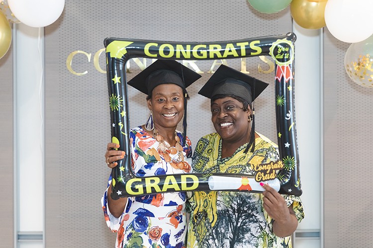 Two women dressed in colorful blouses and a black cap with tassel smile while holding a frame that reads congrat grad
