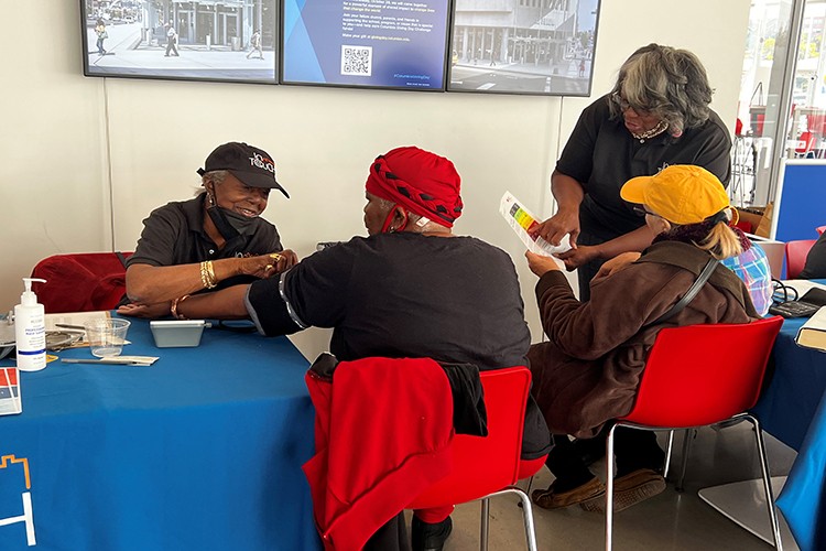 A seated woman in a red headwrap receives Blood Pressure check by a seated female InTOuch member.  Closeby another InTOuch member stands next to a seated woman wearing a yellow ball cap in review of a chart.