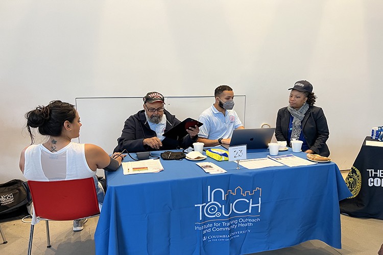 Members of the community are seated around a table with a blue InTOuch banner draped over while in discussion