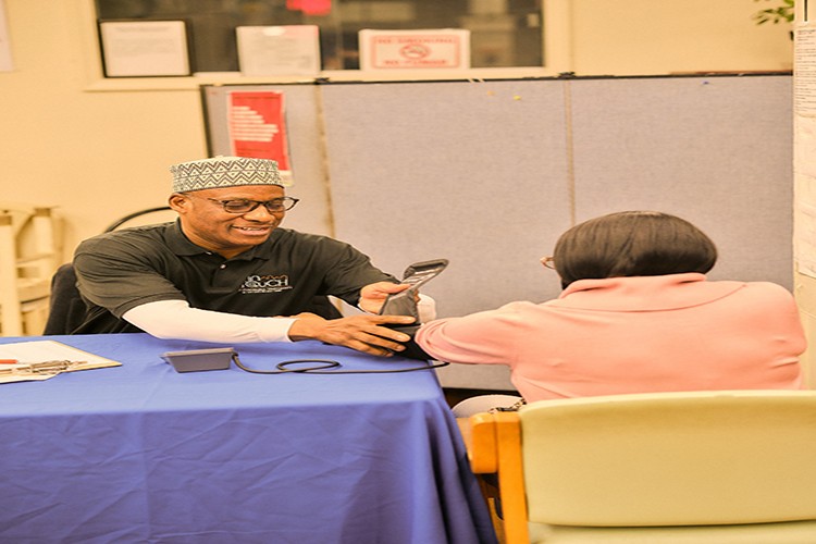 A man dressedin an InTOuch shirt while weairng a kufi and dark rimmed eyeglasses administer blood pressure checking on a seated woman wearing a pink top. 