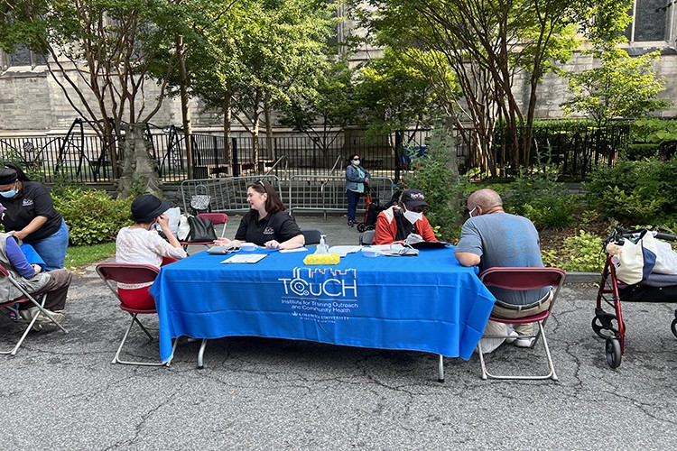 InTOuch members are seated at a table draped in the blue InTOuch banner while speaking with community members during a health screening event