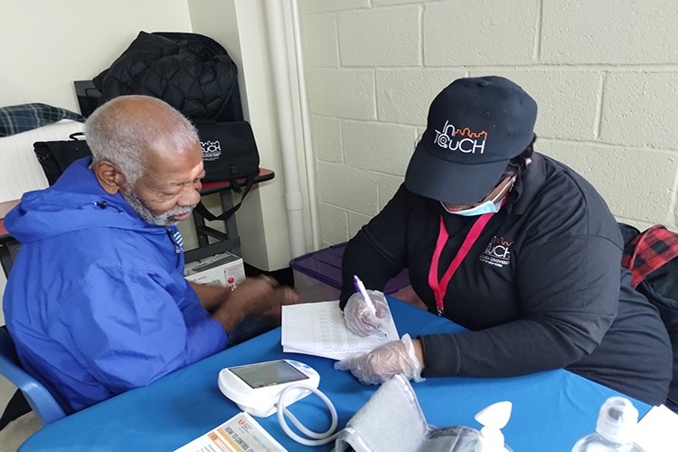 A woman dressed in InTOuch cap and shirt checks an elderly man's blood pressure while noting details on paper.