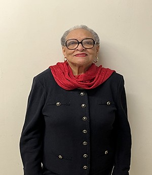 A woman dressed in dark rimmed eyeglasses wears a red shawl draped around her neck wears a dark blazer with gold and black buttons.  She smiles into camera. 