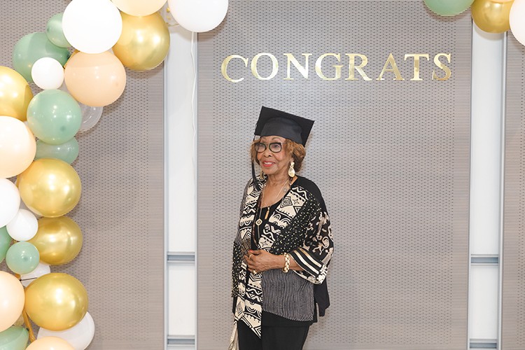 A woman dressed in a black and gold trimmed blouse and dark rimmed eyeglasses wears a black graduation cap while standing underneath a congrats signage and a colorful array of balloons adorn the wall