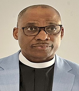 A man wears dark rimmed eyeglass, light grey blazer while wearing a white clerical collar and dark shirt. 