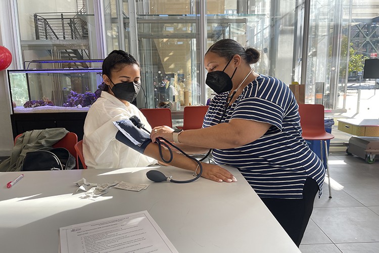 Two women wearing face masks demonstrate blood pressure checking as one woman stands to place the cuff around the arm of a seated womanin a white sweater