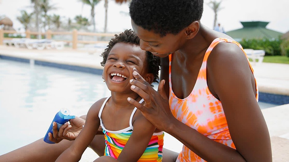 A mother applies sunscreen to her son's face near a pool.