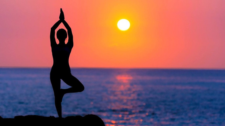 Person stands on a rocky beach with their hands held above their head in a yoga pose as the sun sets over the horizon.