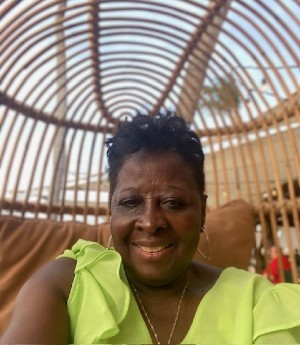 Woman wears a neon lime green short sleeved blouse while seated in a bamboo chair.  She wears a gold chain, earrings and smiles into camera. 