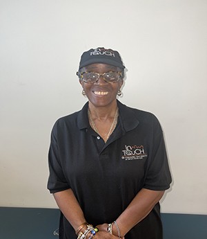 Woman stands in front of a beige wall while smiling into camera.  She wears a dark InTOuch cap and matching polo shirt.  She wears a tortoise shell framed eyeglasses with silver jewelry on her neck and earrings with muticolored bracelets. 