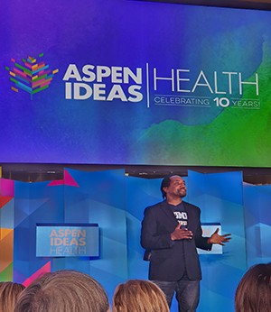 A man stands on stage in front of a projected slide that reads "Aspen Ideas, Health Celebrating 10 Years".  He wears a dark blazer jacket and dark jeans while gestering with open hands to audience.